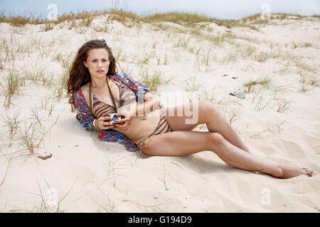 Inspiration captured on wild and wonderful beaches. Bohemian young woman with retro photo camera laying on a white beach Stock Photo