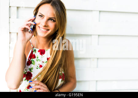 Beautiful young happy woman smiling during a phone call while leaning against a white wall