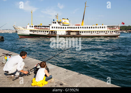 Father and son fishing on quayside, and passenger ferry arriving, in the Bosphorus Strait, Istanbul, Turkey Stock Photo