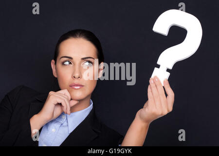 Beautiful businesswoman with a thoughtful expression holding a white question mark in her hand against a black studio background Stock Photo