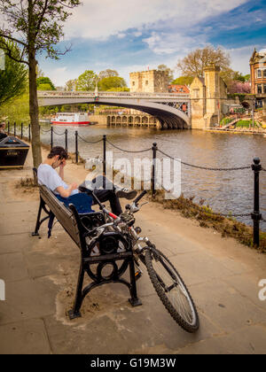 Cyclist sitting on bench checking phone with Lendal Bridge in background, York, UK. Stock Photo