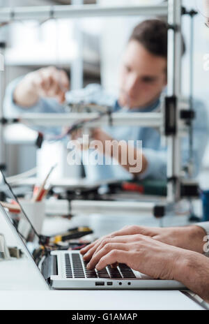 Engineering students working in the lab, a student is adjusting a 3D printer's components, the other one on foreground is using Stock Photo