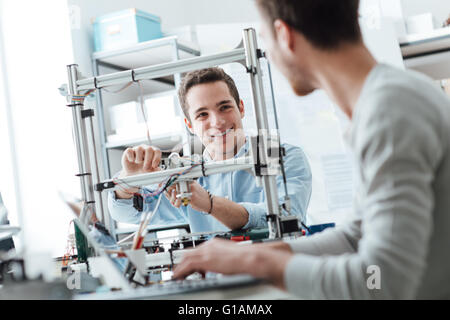 Engineering students working in the lab, a student is adjusting a 3D printer's components, the other one on foreground is using Stock Photo