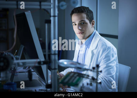 Young researcher in the lab, wearing a lab coat and working with a computer, 3D printer on foreground, he is looking at camera Stock Photo