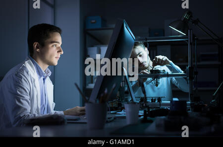 Engineering team working in the lab at night, a student is working with a computer and the other one is adjusting a 3D printer's Stock Photo