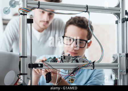 Young engineer in the lab adjusting a 3D printer's components, a student is watching Stock Photo