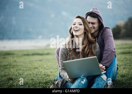 Playful young couple sitting on the grass outdoors, hugging and social networking with a laptop, love and relationships concept Stock Photo