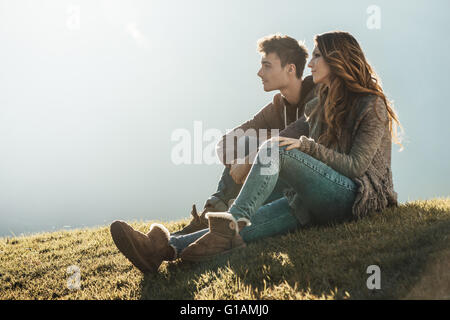 Smiling young couple sitting on grass during a sunny day, looking away and dreaming about their future Stock Photo