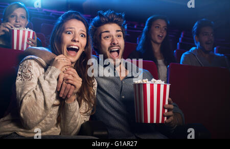 Young scared couple at the cinema watching an horror movie and screaming, she is holding her boyfriend's hand Stock Photo