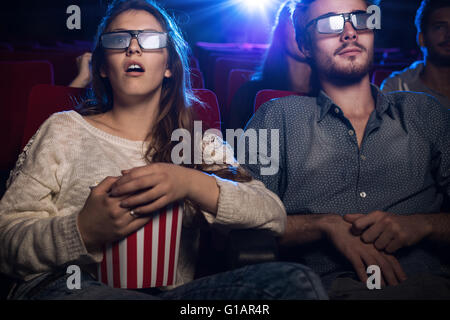 Young teenagers at the cinema wearing glasses and watching a 3d movie, a girl is eating popcorn, entertainment and movies concep Stock Photo
