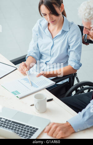 Young business woman on wheelchair working at office desk and checking paperwork with her male colleague, disability Stock Photo