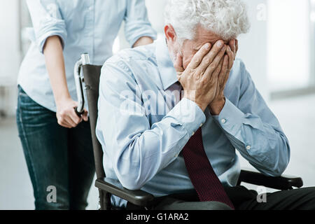 Sad depressed man in wheelchair with head in hands, a woman is taking care of him Stock Photo