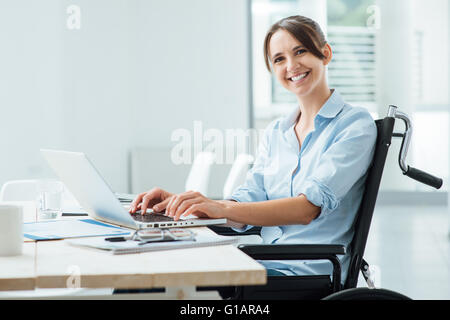 Confident happy businesswoman in wheelchair working at office desk and using a laptop, she is smiling at camera, disability over Stock Photo