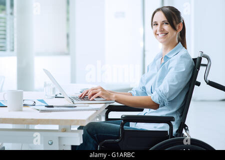Confident happy businesswoman in wheelchair working at office desk and using a laptop, she is smiling at camera, disability over Stock Photo