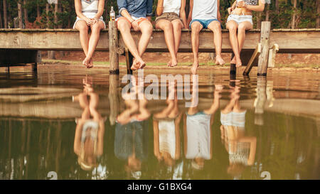 Legs of young people sitting on the edge of a jetty hanging down to the water. Group of friends hanging out at the lake. Stock Photo