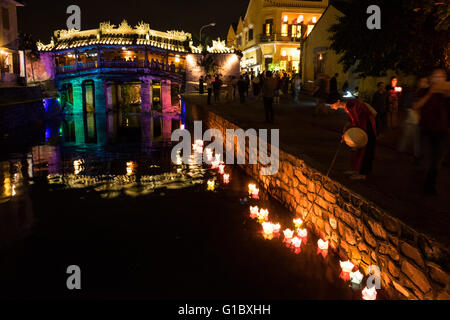 A woman dressed in a traditional vietnamese Ao Dai places a candle into the water next to the historical Japanese Covered Bridge in Hoi An, Vietnam. Stock Photo