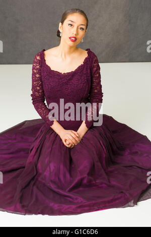 Beautiful young woman in vintage purple dress, sitting on white floor with hands folded in lap. Stock Photo