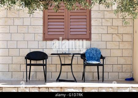Table with two chairs in front of house wall with brown closed shutters Stock Photo