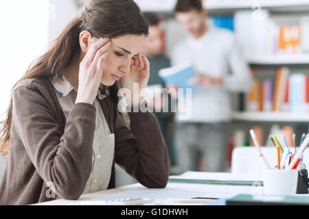 Young female student with headache, she is sitting at desk and touching her head Stock Photo