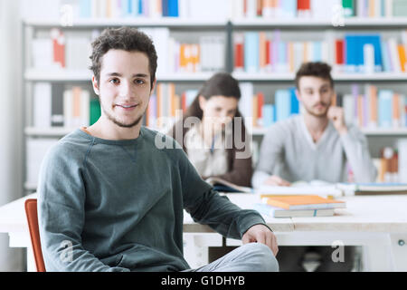 Young male students at the library smiling at camera, his schoolmates are sitting at desk on background Stock Photo