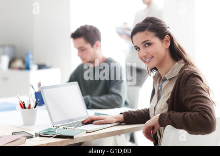 College students in the class, a girl is smiling at camera and using a laptop on foreground Stock Photo