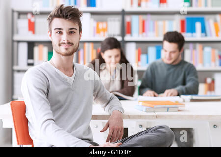 Young male students at the library smiling at camera, his schoolmates are sitting at desk on background Stock Photo