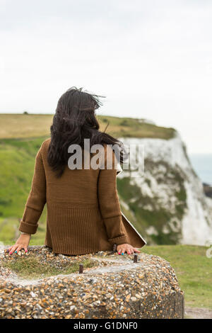 Back of a young woman sitting on a rock and looking at the sea. The White Cliffs of Dover, Dover, Kent, UK Stock Photo