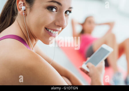 Young smiling woman at the gym having a break and listening to music using a smart phone and earphones, fitness and youth concep Stock Photo