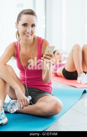 Young smiling woman at the gym having a break and listening to music using a smart phone and earphones, fitness and youth concep Stock Photo