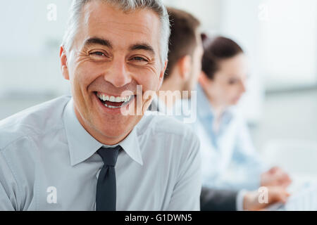 Confident professional businessman smiling at camera, office and business team working on background, selective focus Stock Photo