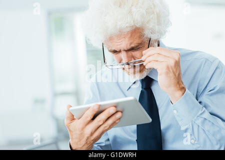 Businessman having eyesight problems, he is using a tablet and adjusting his glasses, office interior on background