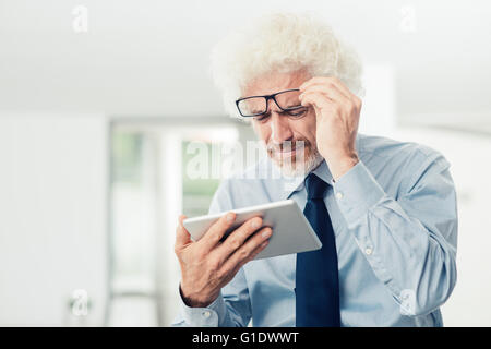 Businessman having eyesight problems, he is using a tablet and adjusting his glasses, office interior on background Stock Photo