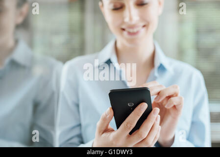 Beautiful smiling young woman using a smart phone, leaning on a window and reflecting on glass Stock Photo