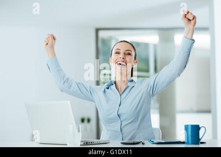 Successful cheerful businesswoman with fists raised sitting at office desk, achievement and satisfaction concept Stock Photo