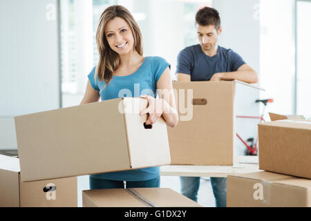 Young happy couple moving in their new house and unpacking boxes, she is carrying a carton box and smiling at camera Stock Photo