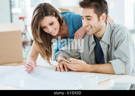 Young couple planning their new dream house, he is sitting at desk and she is drawing on a project, carton boxes on background Stock Photo