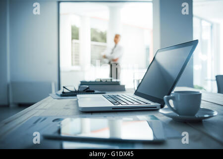 Businessman standing with arms crossed in his office, desktop with laptop on foreground, selective focus Stock Photo