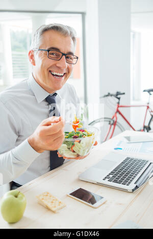 Happy smiling businessman having an healthy salad for lunch, he is sitting at office desk and looking at camera Stock Photo