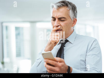 Confused businessman with hand on chin having troubles using a smart phone Stock Photo