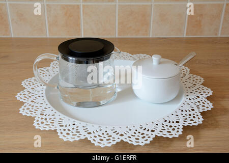 Glass tea kettle and sugar bowl with spoon on white metal round tray on brown wooden table in kitchen Stock Photo