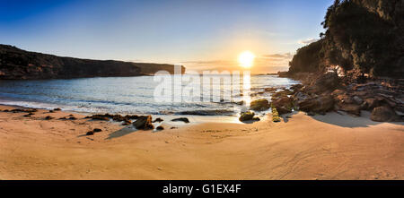 WIde morning panorama of Wattamola beach clean sand and low tide rocks in Royal National park, Australia. Bright rising sun over Stock Photo