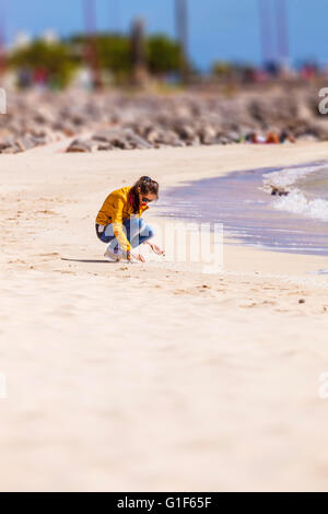 MODEL RELEASED. Young woman crouching on the beach collecting shells. Stock Photo