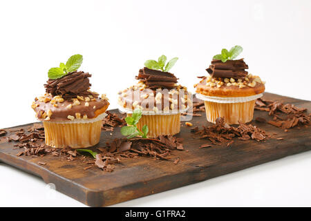 three chocolate muffins with chopped nuts on wooden cutting board Stock Photo