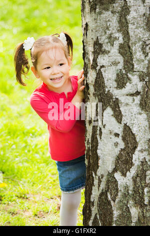 Child playing hide and seek outdoors in park. Beautiful little girl hiding behind huge tree on summer or spring sunny day Stock Photo