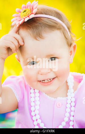 Close up portrait of charming smiley face of one year old small girl dressed in cute pink dress, white necklace and pink flower Stock Photo