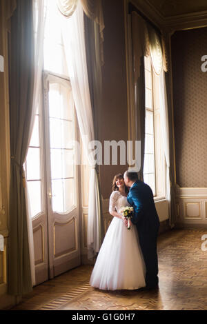 Handsome husband kissing and embracing his wife at gorgeous window on the background of rich interior in old building Stock Photo