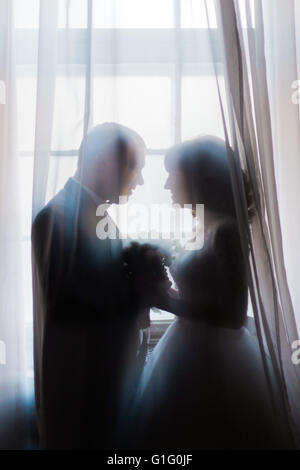 Silhouette of bride and groom embracing holding bouquet against the window with curtains Stock Photo