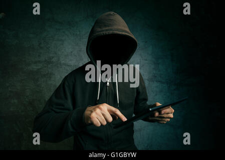 Faceless hooded unidentifiable male person using tablet computer, internet hacker and online security concept. Stock Photo