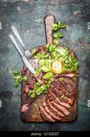 Sliced medium rare grilled beef barbecue steak served with fresh green salad and cutlery on rustic  cutting board , top view.  M Stock Photo