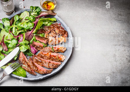 Sliced grilled beef steak with green leaves salad on rustic plate with cutlery. Medium rare barbecue steak and healthy salad on Stock Photo